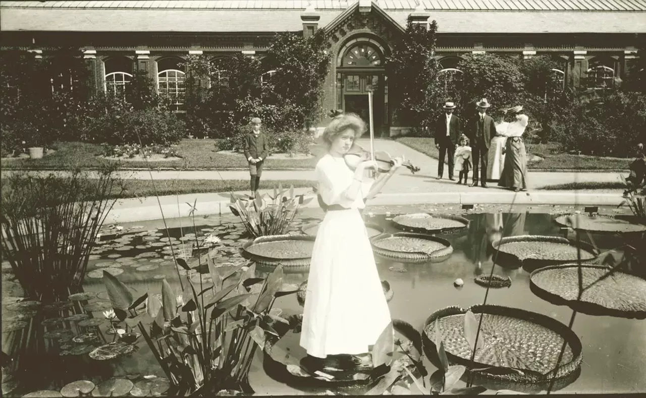 A young girl plays a violin while standing on lily pad in front of Linnean House at Shaw's Garden (now the Missouri Botanical Garden), ca. 1900&#150;1910.