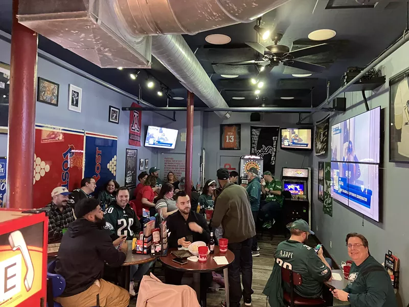 People in Eagles green sit around tables in a bar watching TV.